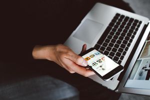 Hand holding a smart phone open to apps screen. Hand and phone are being held over an open laptop
