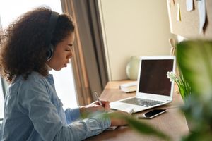 Person writing in notebook while listening to headphones. Open laptop and phone are also on the desk.