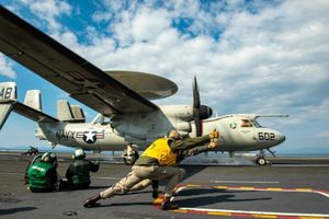 Aircraft launching from carrier deck, with crew in safety gear signaling takeoff.