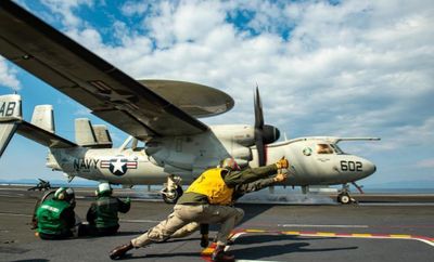 Aircraft launching from carrier deck, with crew in safety gear signaling takeoff.