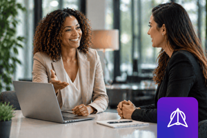 Two women having a professional conversation at a table, one gesturing toward an open laptop.