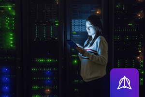 A woman standing in a darkened data center, looking at an tablet, with glowing green, blue and orange lights visible from the racks behind her.