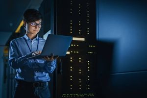 IT professional working on a laptop in a dimly lit data center aisle with server racks illuminated in the background.